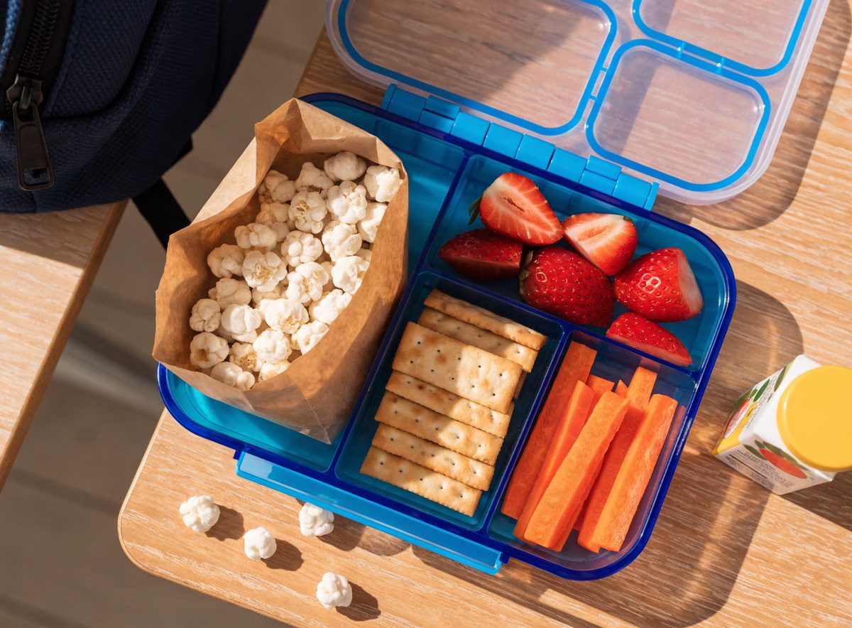 Open children's lunchbox packed with popped sorghum in a kraft bag, strawberries, crackers, and carrot sticks