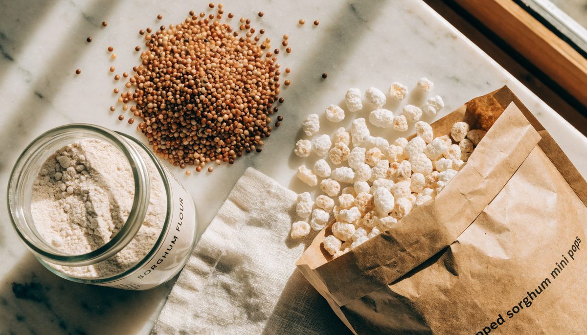 Sorghum flour in a jar, raw sorghum kernels, and a bag of popped sorghum mini pops on a marble countertop