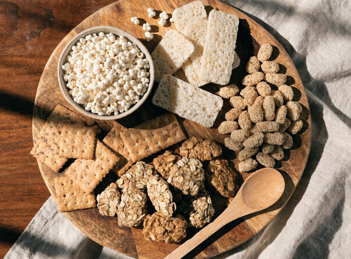 Overhead arrangement of whole grain snacks on a wooden board: popped sorghum, crackers, oat clusters, and rice cakes
